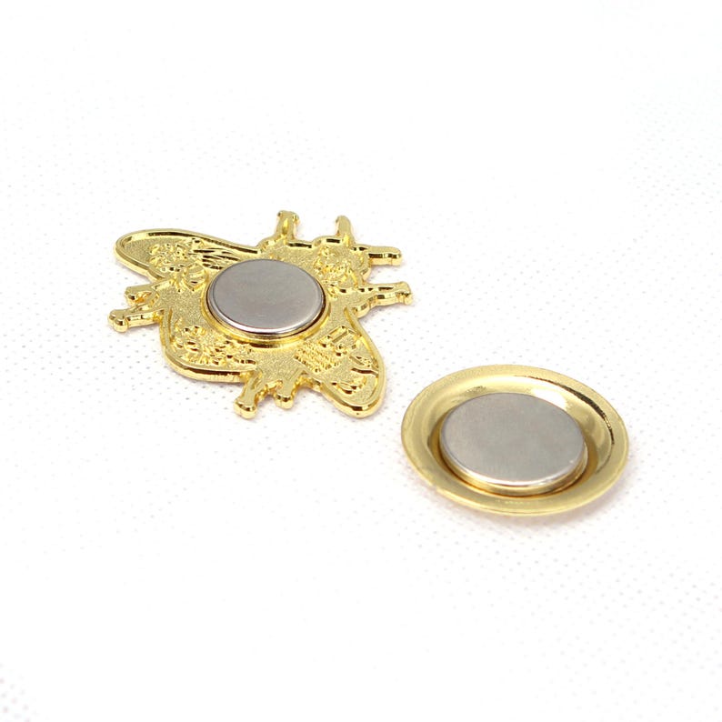 May include: A gold-colored bee-shaped magnet with intricate detailing, next to a matching round magnet. The magnets have a silver-colored center and are set against a white background. The bee magnet is approximately 1.5 inches wide.