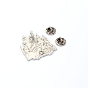 May include: A silver-colored pin featuring a detailed castle design with text. Two additional pin backs are included. The pin is likely made of metal and has a shiny finish. The design is intricate, showing architectural details.