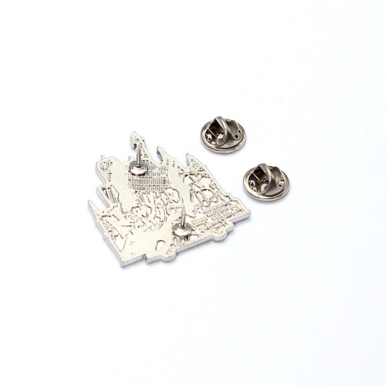 May include: A silver-colored pin featuring a detailed castle design with text. Two additional pin backs are included. The pin is likely made of metal and has a shiny finish. The design is intricate, showing architectural details.