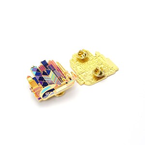 May include: Two enamel pins featuring a colorful cityscape design. The pins showcase a skyline with buildings in shades of pink, purple, blue, and gold. One pin is face up, while the other reveals the gold-colored back with a pin clasp.