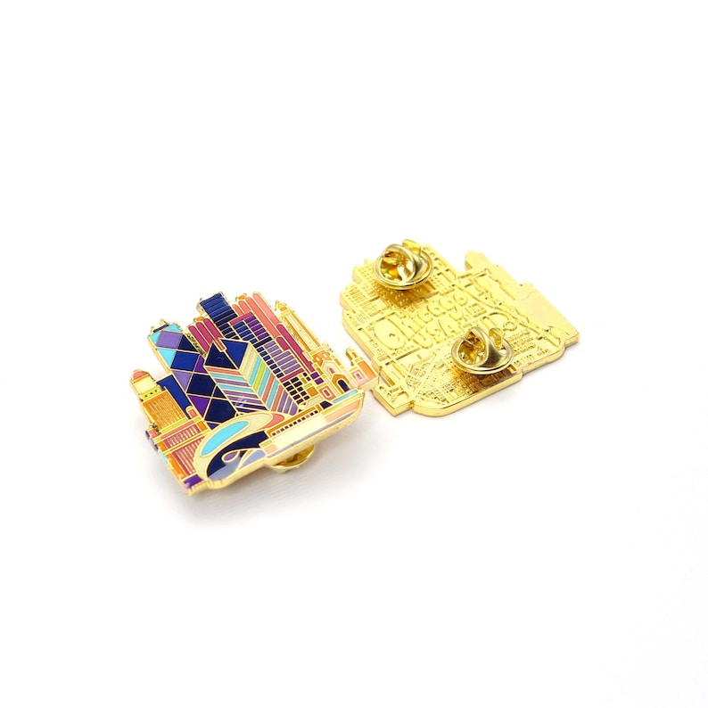 May include: Two enamel pins featuring a colorful cityscape design. The pins showcase a skyline with buildings in shades of pink, purple, blue, and gold. One pin is face up, while the other reveals the gold-colored back with a pin clasp.