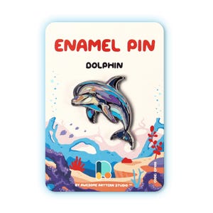 May include: An enamel pin shaped like a dolphin with a multi-colored abstract pattern of blue, purple, and white. The pin is attached to a rectangular display card featuring the text ENAMEL PIN, DOLPHIN, and BY AWESOME PATTERN STUDIO.
