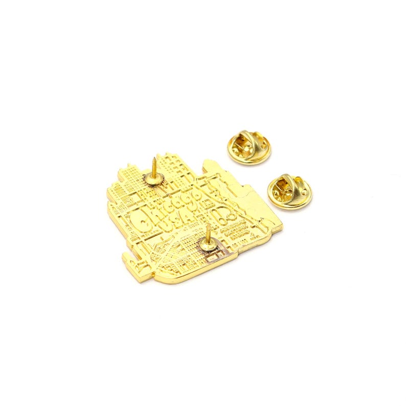 May include: A gold-colored pin with the text "Chicago USA" and a map design. The pin has two pin backs and a rectangular shape. The pin is likely made of metal and has a shiny finish.