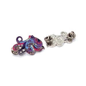 May include: Two octopus-shaped enamel pins. One pin features a colorful design with pink, purple, blue, and red details. The other pin shows the back with a pin clasp. The pins are on a white background.