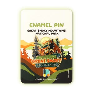 May include: Enamel pin featuring a cartoon illustration of a bear and a cub in front of a mountain range with a sunset. The text "Great Smoky Mountains National Park" is written in a gold banner below the image. The pin is on a white card with the text "Enamel Pin" and "By Awesome Pattern Studio TM" printed on it.