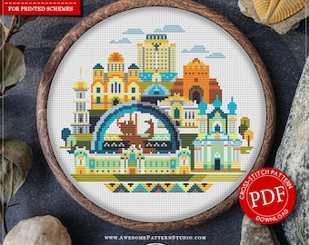 May include: A cross-stitch pattern featuring a colorful cityscape design, displayed in a wooden embroidery hoop. The pattern includes buildings and architectural elements in shades of blue, orange, yellow, and green. The text "COUNTED CROSS STITCH PATTERN" and "PDF DOWNLOAD" are visible.
