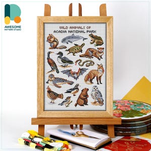 May include: A framed cross-stitch sampler titled "Wild Animals of Acadia National Park" featuring various wildlife illustrations. The sampler is displayed on a wooden easel, with embroidery supplies in the foreground. The Awesome Pattern Studio logo is in the upper left corner.