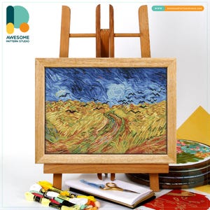 Puede incluir: Un punto de cruz enmarcado de un paisaje con un camino sinuoso, un campo de trigo y un cielo estrellado, expuesto en un caballete de madera. La obra de arte está en un marco de madera. Hilo de bordar, tijeras y un cuaderno están en primer plano.