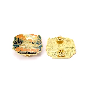 May include: Two Glacier National Park enamel pins. One pin features a landscape with mountains, trees, and the text "Glacier National Park" in orange and blue. The other pin is gold-colored with a mountain and tree design.