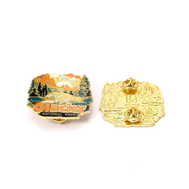 May include: Two Glacier National Park enamel pins. One pin features a landscape with mountains, trees, and the text "Glacier National Park" in orange and blue. The other pin is gold-colored with a mountain and tree design.
