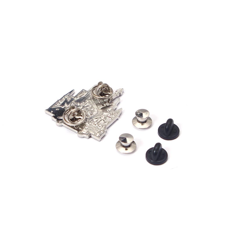 May include: A silver-colored metal pin with a detailed architectural design, featuring two pin backs. Three silver pin clutches and two black rubber clutches are also visible, all against a white background.