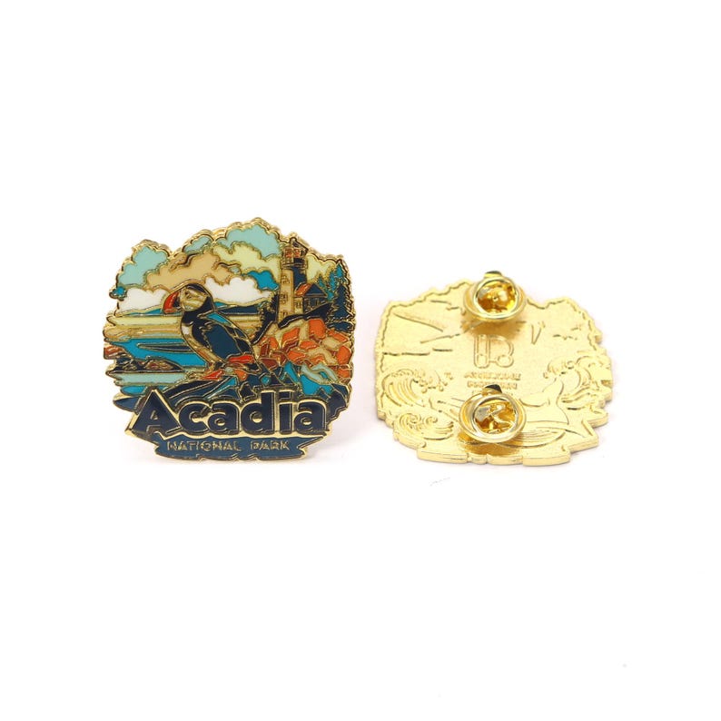 K&ouml;nnte beinhalten: Ein farbenfroher Emaille-Pin mit einem Papageientaucher, einem Leuchtturm und einer Meereslandschaft. Der Pin tr&auml;gt die Aufschrift "Acadia National Park" in Marineblau. Die R&uuml;ckseite ist goldfarben mit zwei Nadelverschl&uuml;ssen.