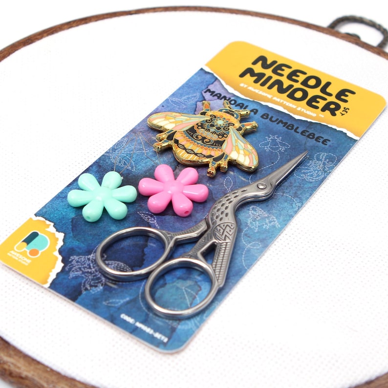 May include: A craft kit featuring a "Needle Minder" set with a decorative bumblebee pin, two flower-shaped stitch markers, and a pair of silver embroidery scissors. The items are on a blue patterned card, set against a white background.