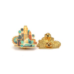 May include: A gold-colored enamel pin featuring a cityscape design with a Ferris wheel, palm trees, and various buildings. The pin has a detailed front and back, showcasing the pin's construction and pin backs.
