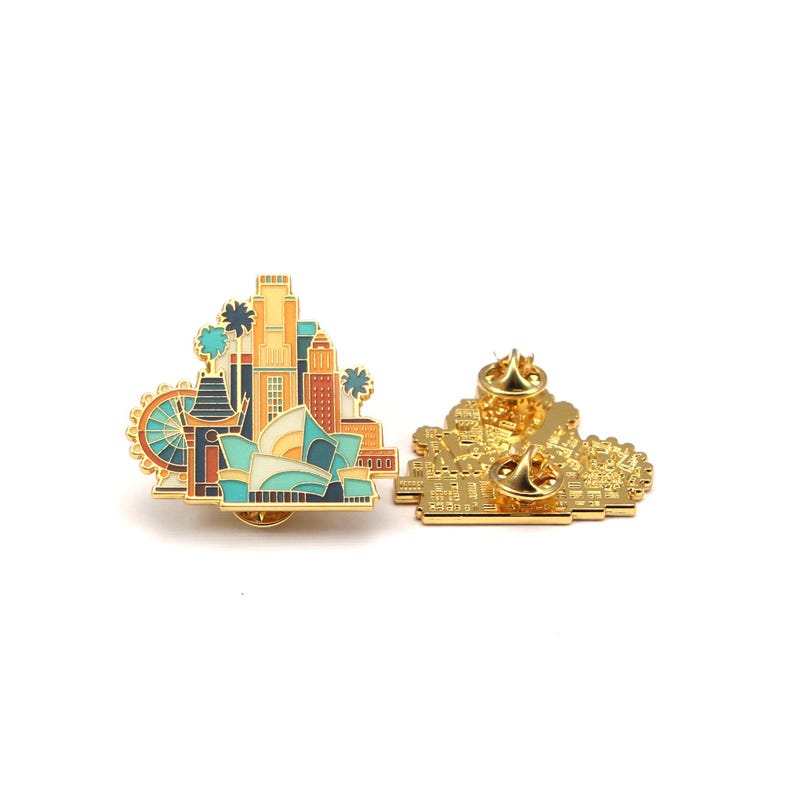 May include: A gold-colored enamel pin featuring a cityscape design with a Ferris wheel, palm trees, and various buildings. The pin has a detailed front and back, showcasing the pin's construction and pin backs.