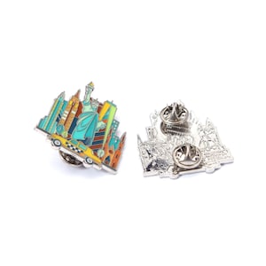 May include: Enamel pin featuring a colorful New York City skyline, including the Statue of Liberty and a yellow taxi cab. The pin has a silver backing with two pin posts for secure attachment. A souvenir or collectible item.