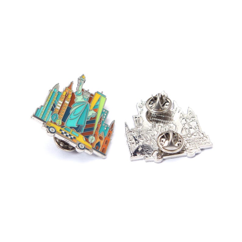 May include: Enamel pin featuring a colorful New York City skyline, including the Statue of Liberty and a yellow taxi cab. The pin has a silver backing with two pin posts for secure attachment. A souvenir or collectible item.
