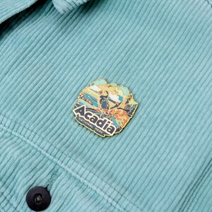 K&ouml;nnte beinhalten: Ein goldfarbener Emaille-Pin mit einer farbenfrohen Illustration des Acadia Nationalparks mit einem Papageientaucher und dem Text "Acadia National Park". Der Pin ist an einer tealfarbenen Cordjacke befestigt.