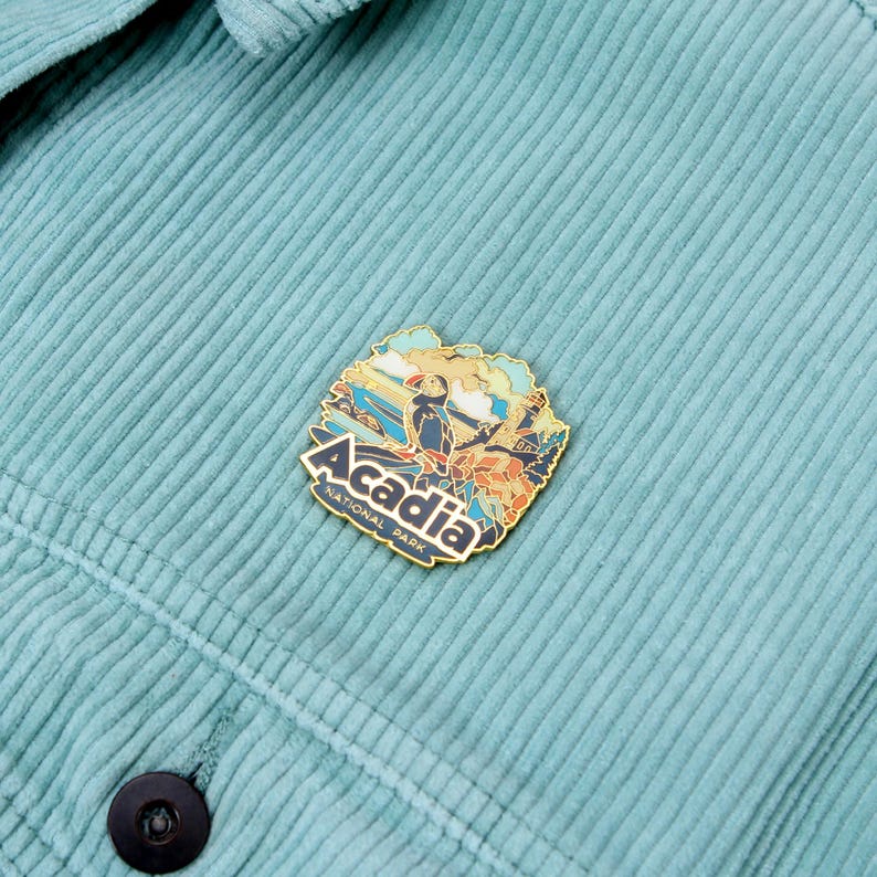K&ouml;nnte beinhalten: Ein goldfarbener Emaille-Pin mit einer farbenfrohen Illustration des Acadia Nationalparks mit einem Papageientaucher und dem Text "Acadia National Park". Der Pin ist an einer tealfarbenen Cordjacke befestigt.