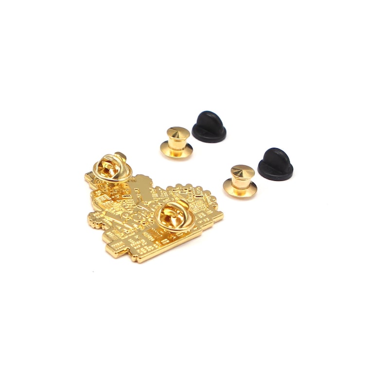 May include: A gold-colored pin with a detailed design, featuring a pin back and two black rubber clutches. The pin has a complex, pixelated pattern. Two additional gold pin backs are also visible.