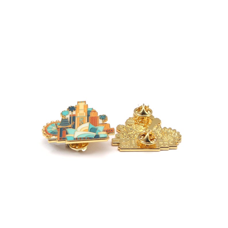 May include: Two gold-colored enamel pins. One pin features a colorful cityscape design with buildings, a Ferris wheel, and palm trees. The other pin shows the back of the cityscape pin with two pin backs.