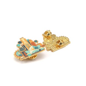 May include: Two gold-colored enamel pins. One pin features a colorful cityscape design with a Ferris wheel, buildings, and palm trees. The other pin shows the back of the pin with a gold-colored pin back.