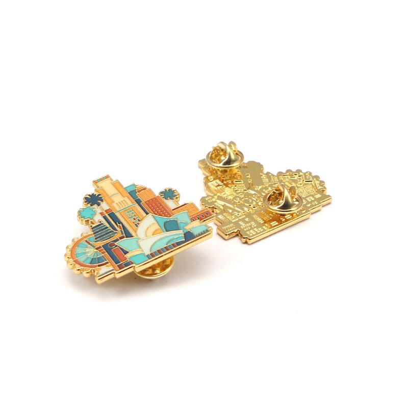 May include: Two gold-colored enamel pins. One pin features a colorful cityscape design with a Ferris wheel, buildings, and palm trees. The other pin shows the back of the pin with a gold-colored pin back.
