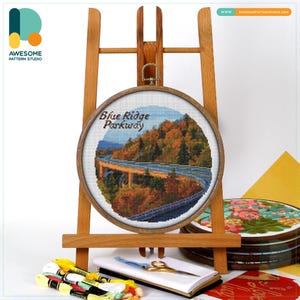 Könnte beinhalten: Ein Kreuzstich-Stickrahmen mit einer Landschaft der Blue Ridge Parkway, ausgestellt auf einer Holzstaffelei. Die Stickerei zeigt eine Brücke und Herbstlaub. Nähzubehör, einschließlich Faden und Schere, ist ebenfalls sichtbar. Der Rahmen hat etwa 20 cm Durchmesser.