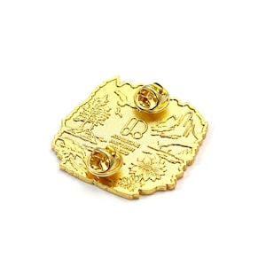 May include: A gold-colored enamel pin with a detailed landscape design. The pin features a tree, mountains, and a flower, with the text "AWESOME PATTERN STUDIO". The pin has two pin backs for secure attachment.