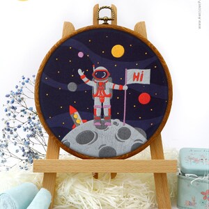 Pode incluir: Arte de bordado em bastidor com um astronauta na lua com um foguete e planetas contra um céu estrelado azul escuro. O astronauta segura uma bandeira que diz "Hi". A obra de arte é exibida em um cavalete de madeira.