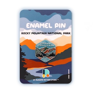 May include: An enamel pin featuring a Rocky Mountain National Park design. The pin is a hexagon shape with a detailed landscape illustration in shades of blue, green, and brown. The pin is mounted on a card with text that reads "ENAMEL PIN" and "ROCKY MOUNTAIN NATIONAL PARK."