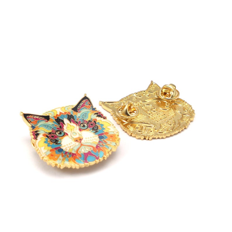 Puede incluir: Un pin decorativo con forma de cara de gato con un reverso de metal dorado. El pin presenta un dise&ntilde;o detallado con una variedad de colores, incluyendo azul, amarillo, naranja y rosa. El pin mide aproximadamente 2,5 cm de di&aacute;metro.