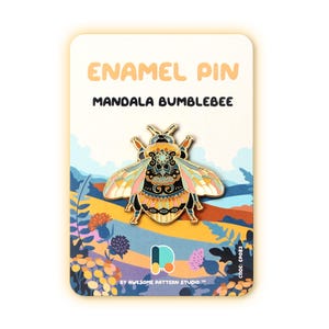 May include: An enamel pin featuring a mandala bumblebee design. The pin has a detailed, colorful pattern with gold accents. The pin is displayed on a card with the text "ENAMEL PIN MANDALA BUMBLEBEE". The card has a landscape illustration with flowers and mountains.
