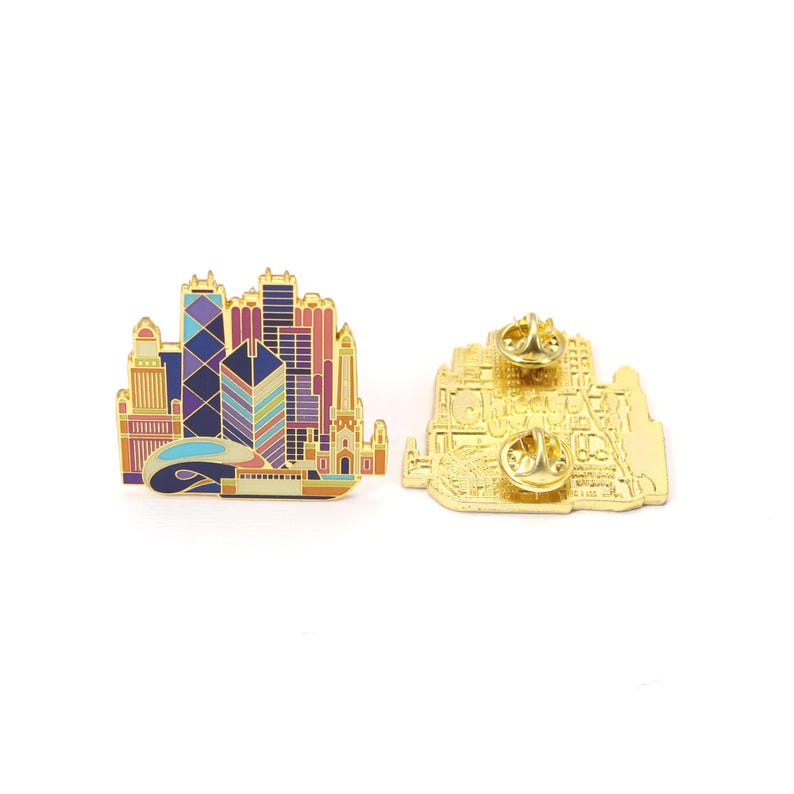 May include: A gold-colored enamel pin featuring a colorful cityscape design. The pin depicts a skyline with various buildings in shades of blue, purple, orange, and pink. The pin has a butterfly clutch backing. The back of the pin is also gold.