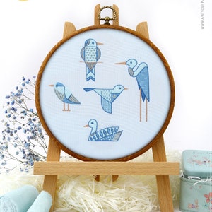 Puede incluir: Un bastidor de bordado con cinco pájaros azules estilizados sobre un fondo azul claro. Los pájaros tienen diseños geométricos y se muestran en un bastidor de madera sobre un caballete de madera. El texto "Awesome Pattern Studio with love" es visible.