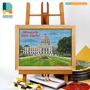 May include: A framed cross-stitch of the Minnesota State Capitol building in Saint Paul, displayed on a wooden easel. The artwork features a detailed depiction of the capitol building, with green grass and a blue sky. Embroidery threads, scissors, and a notebook are in the foreground.