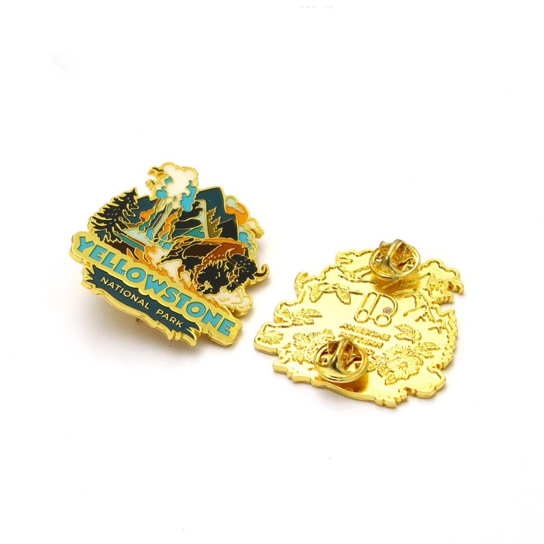 May include: Gold-colored enamel pin featuring a colorful illustration of Yellowstone National Park with the text "Yellowstone National Park". The back of the pin shows a detailed outline of the park.