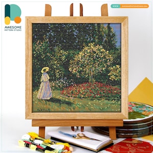May include: Cross-stitch artwork of a woman in a garden, framed in a light wood frame. The scene depicts a woman in a long dress and hat, walking through a garden with trees and flowers. The artwork is displayed on a wooden easel, with embroidery supplies nearby.