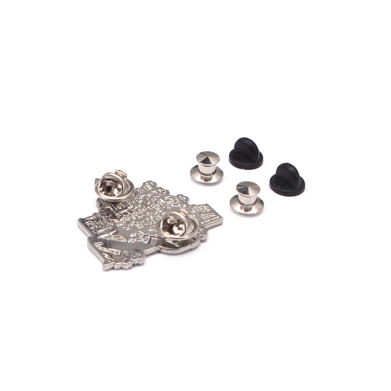May include: A silver-colored metal pin in the shape of a map, with intricate detailing. The pin has two pin backs and is accompanied by three silver pin backs and two black rubber pin keepers. The pin is on a white background.