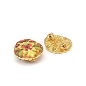 Puede incluir: Dos broches decorativos de esmalte con acabado dorado. Un broche presenta un dise&ntilde;o de le&oacute;n estilizado y colorido con detalles en rojo, naranja, amarillo y verde. El otro broche muestra el reverso dorado con un cierre de alfiler.