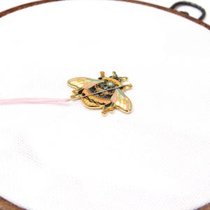 May include: A gold-colored bee-shaped enamel pin with intricate details in black, orange, pink, and blue. The pin is attached to a pink thread, set against a white fabric within a wooden embroidery hoop. The bee pin is a decorative craft item.