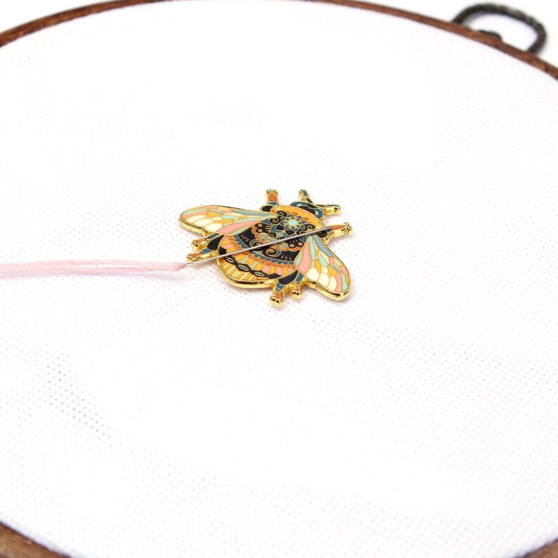 May include: A gold-colored bee-shaped enamel pin with intricate details in black, orange, pink, and blue. The pin is attached to a pink thread, set against a white fabric within a wooden embroidery hoop. The bee pin is a decorative craft item.