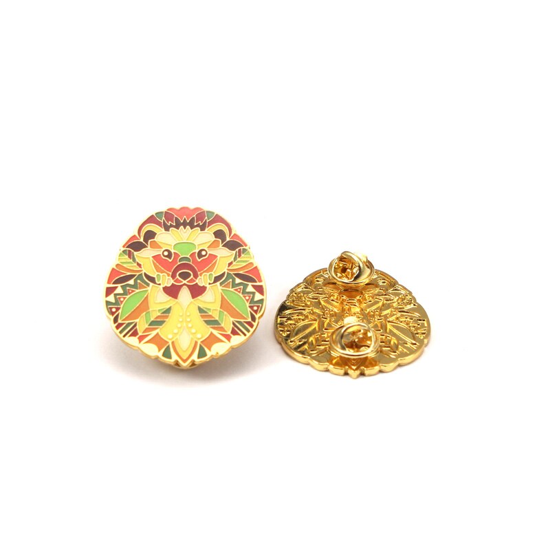 Puede incluir: Un broche de esmalte dorado con forma de erizo con un dise&ntilde;o geom&eacute;trico colorido. El broche presenta tonos de rojo, amarillo, verde y naranja. La parte posterior del broche tambi&eacute;n es dorada y muestra el cierre.