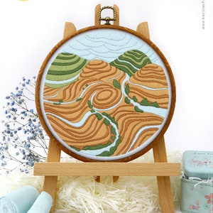 May include: Embroidered landscape art in a wooden hoop, displayed on a small wooden easel. The artwork depicts rolling hills in shades of brown, green, and blue, with a light blue sky. The text "AWESOME Pattern Studio with love" is visible.