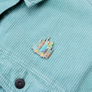 May include: A colorful enamel pin featuring a New York City skyline design, including the Statue of Liberty, skyscrapers, and a yellow taxi cab. The pin is set against a light teal corduroy jacket.