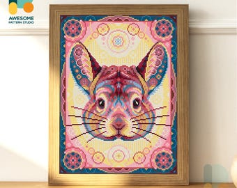 May include: A framed cross-stitch artwork depicting a symmetrical rodent face in vibrant colours. The design incorporates pink, blue, yellow, and red. The frame is light brown wood. The website address "AWESOMEPATTERNSTUDIO.COM" is visible.