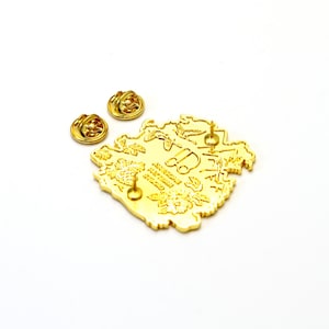 May include: Gold-toned enamel pin featuring a detailed illustration of a floral design with a small car in the center. The pin has a text inscription that reads "AMERICAN DREAM STUDIO".
