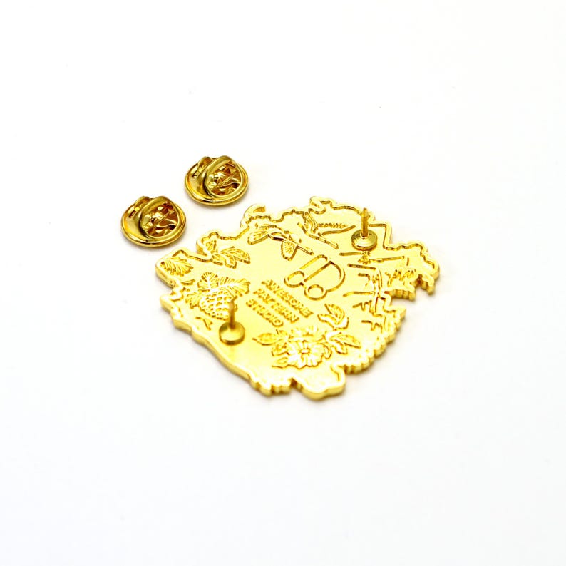 May include: Gold-toned enamel pin featuring a detailed illustration of a floral design with a small car in the center. The pin has a text inscription that reads "AMERICAN DREAM STUDIO".