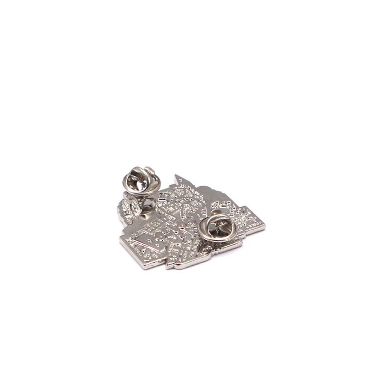 May include: A silver metal pin with a detailed, textured design. The pin has two pin backs for secure attachment. The design appears to be an abstract representation of a city or map. The pin is set against a white background.