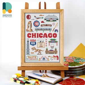 May include: A framed cross-stitch artwork featuring Chicago landmarks, including the skyline, Navy Pier, and Route 66. The word "CHICAGO" is prominently displayed. The artwork is displayed on a wooden easel, with embroidery supplies in the foreground.